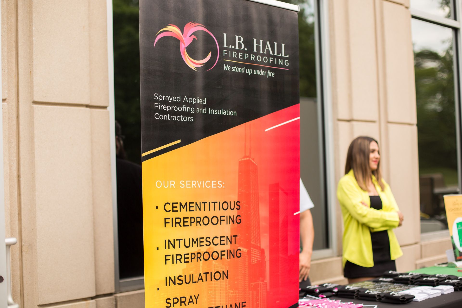 Outdoor booth banner for L.B. Hall, with a woman in a yellow jacket standing beside it.