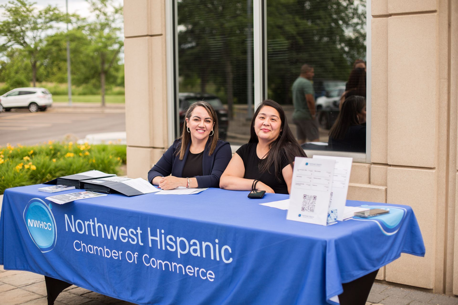 Two women at a Northwest Hispanic Chamber of Commerce booth with a blue tablecloth outdoors