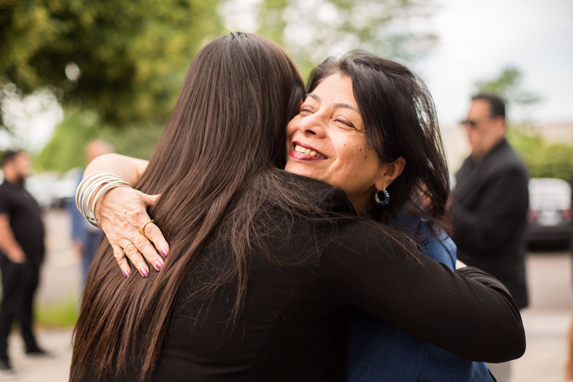 Two people hugging outdoors, one smiling with eyes closed in a warm embrace.