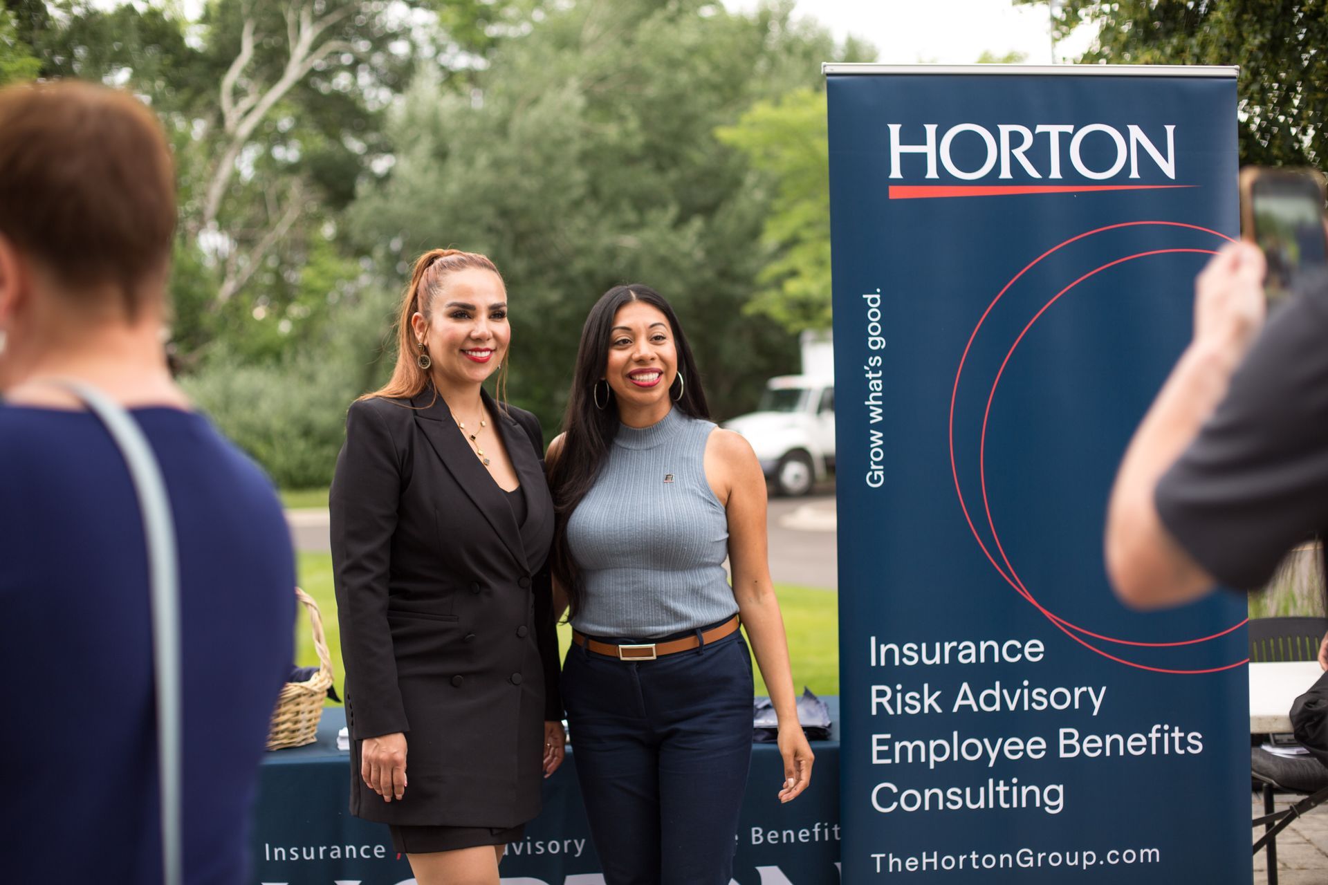 Two women posing beside a HORTON insurance and benefits consulting banner outdoors
