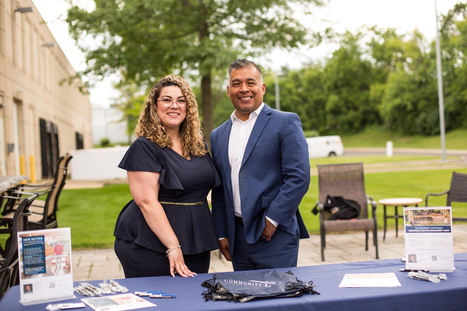 Two people stand behind a table with brochures and lanyards outdoors near a building and lawn.