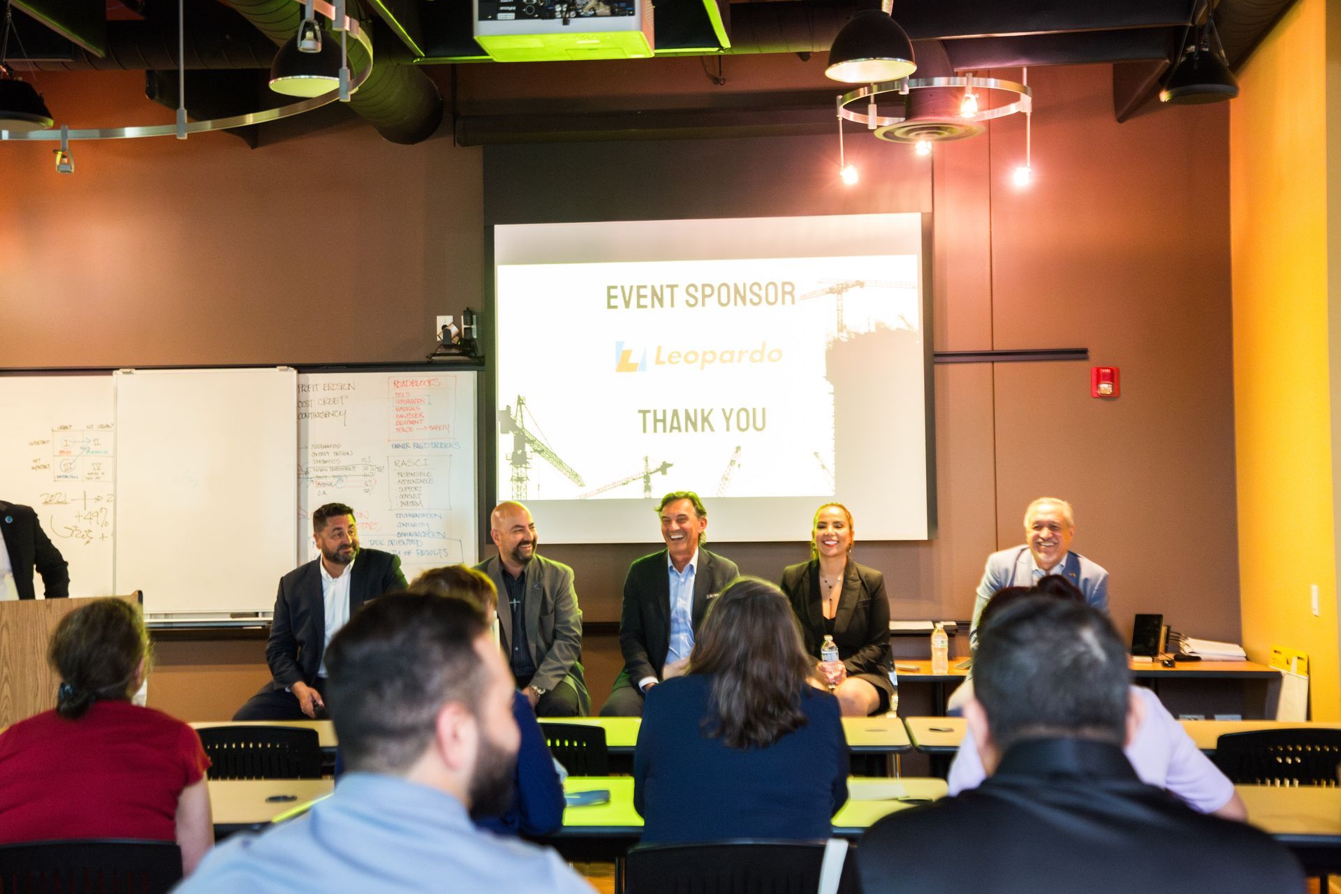 Panel discussion in a conference room, with five speakers seated before an audience and a projected presentation screen.