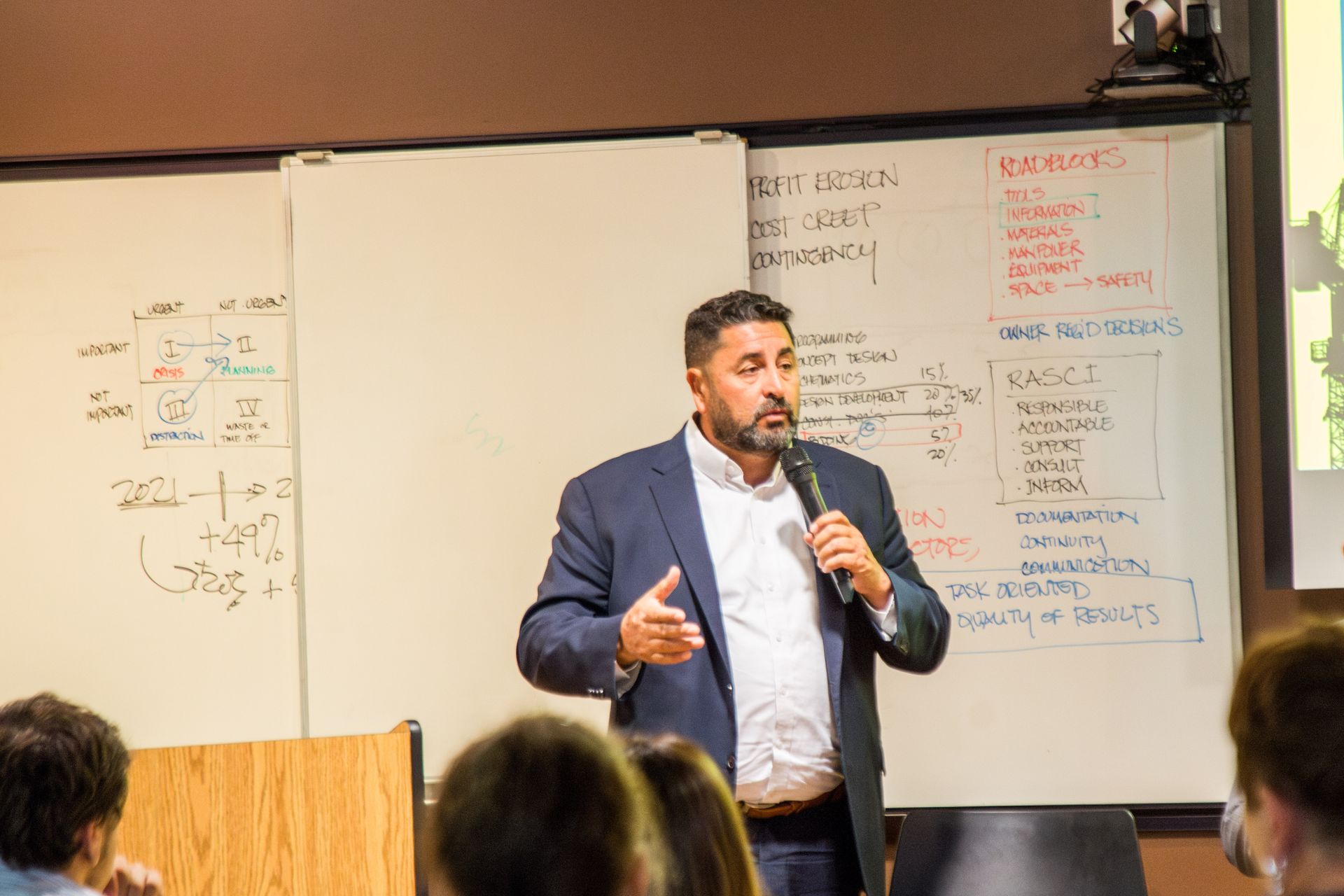Man speaking into a microphone beside a whiteboard in a classroom or meeting room