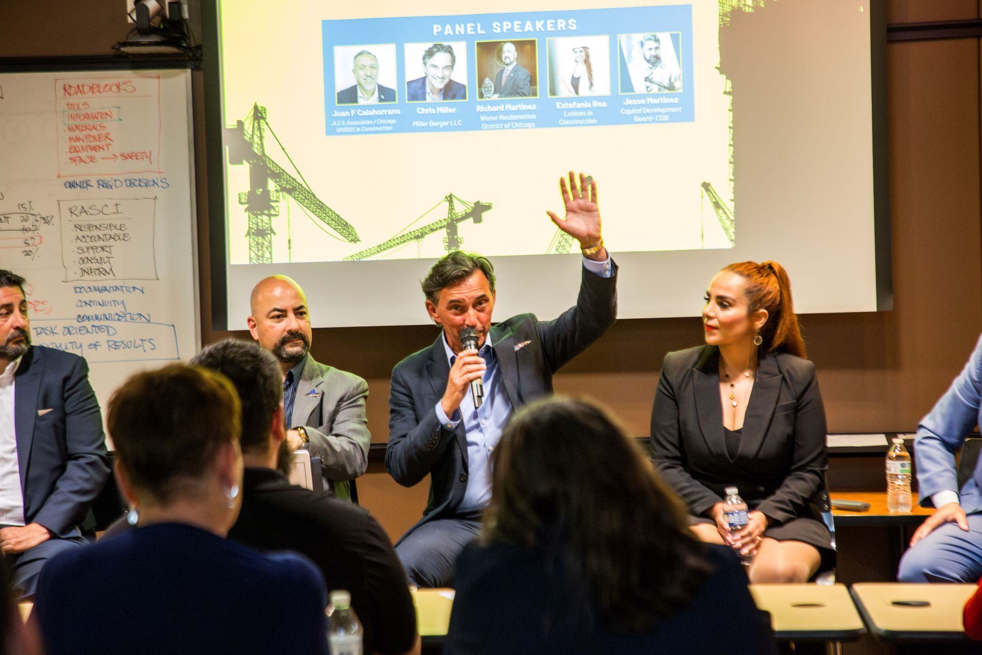 Panel discussion on stage, with one speaker raising a hand and a projected presentation behind them.