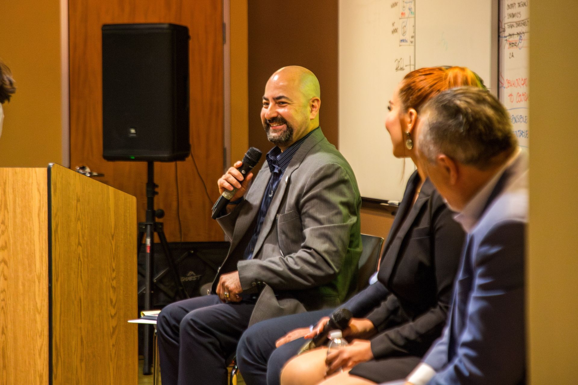 Three panelists seated onstage, one speaking into a microphone, in a conference room.