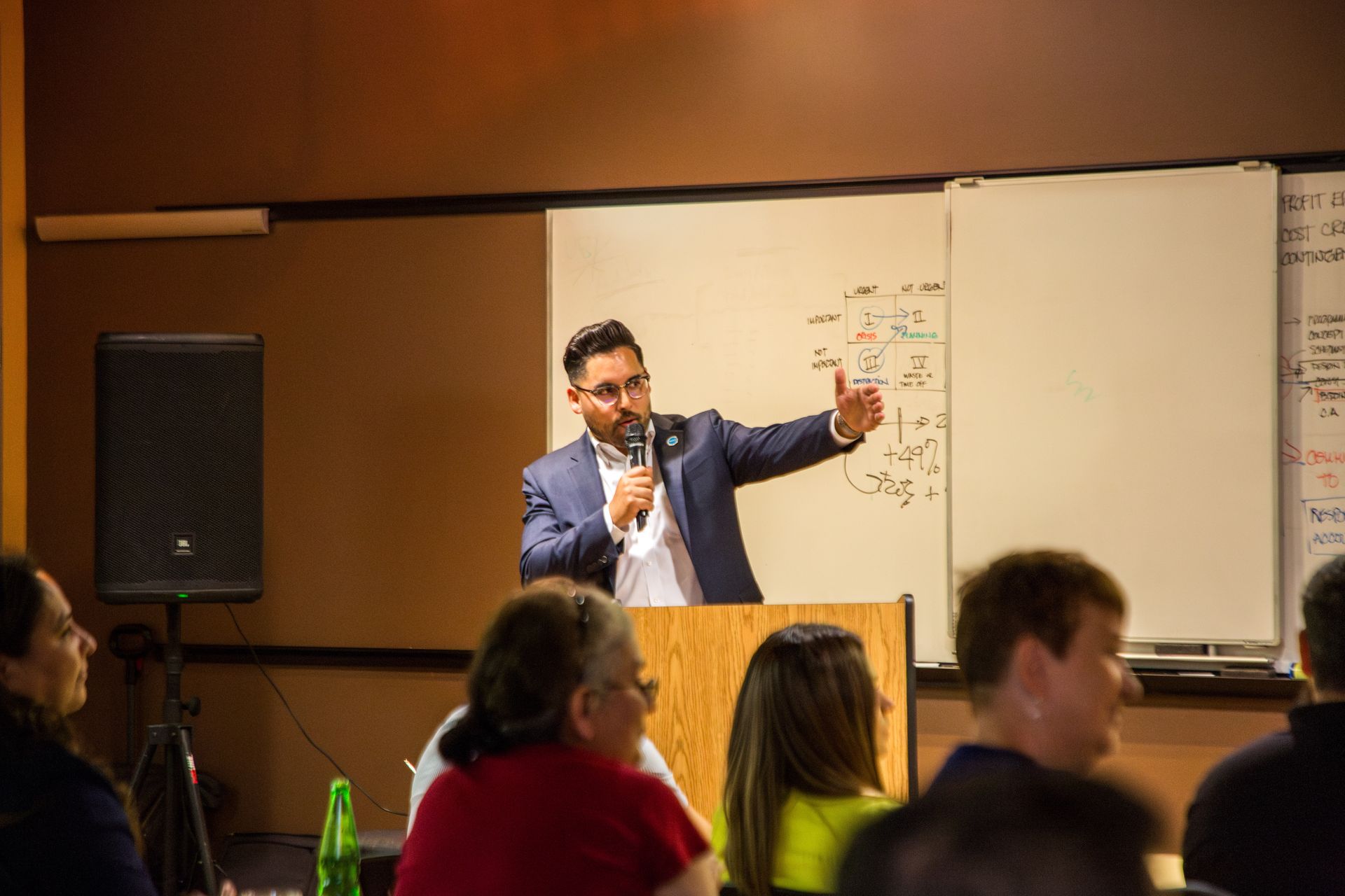 Speaker in a blue suit presents at a podium in a conference room, gesturing toward a whiteboard.