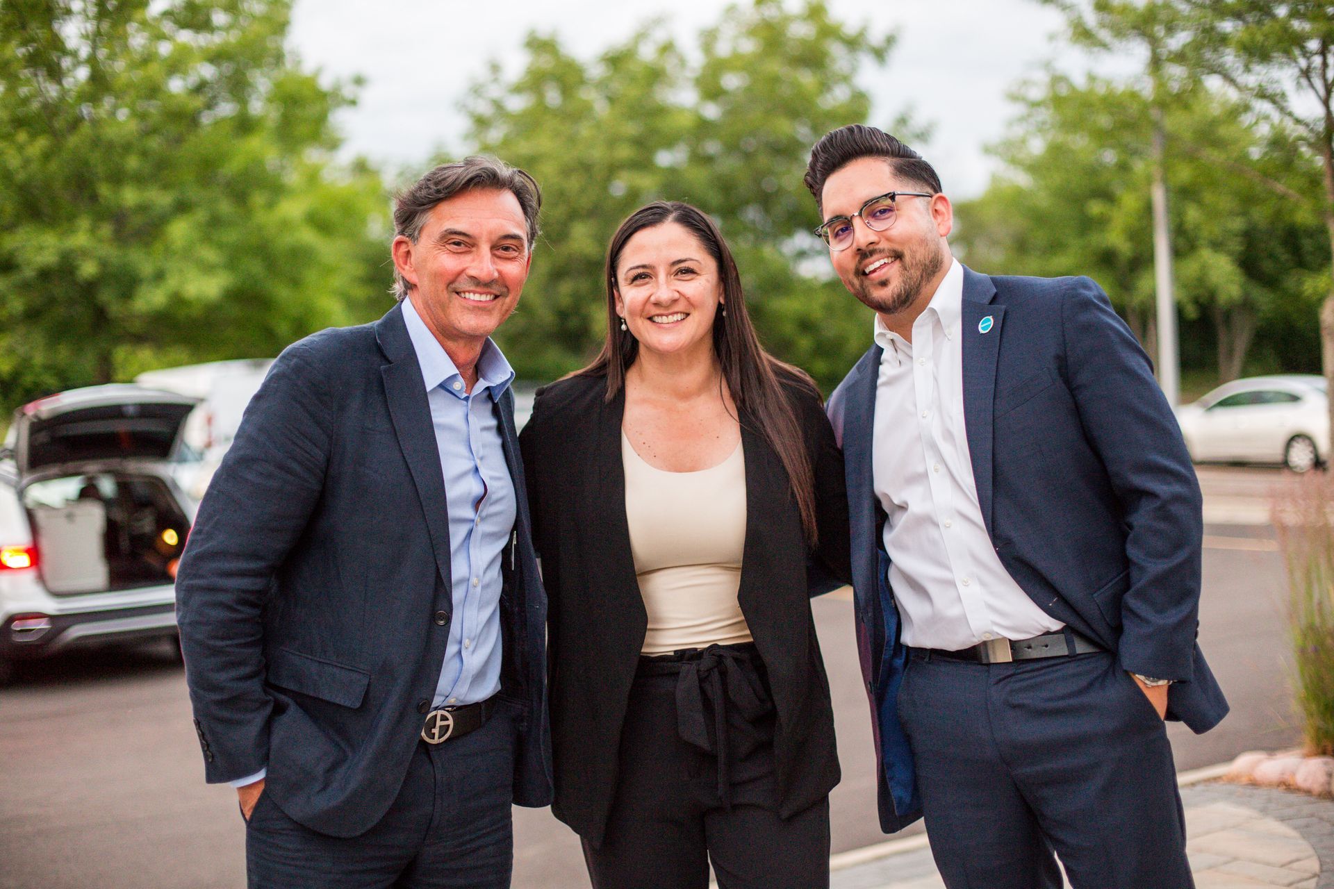 Three people smiling outdoors near parked cars, dressed in business attire.