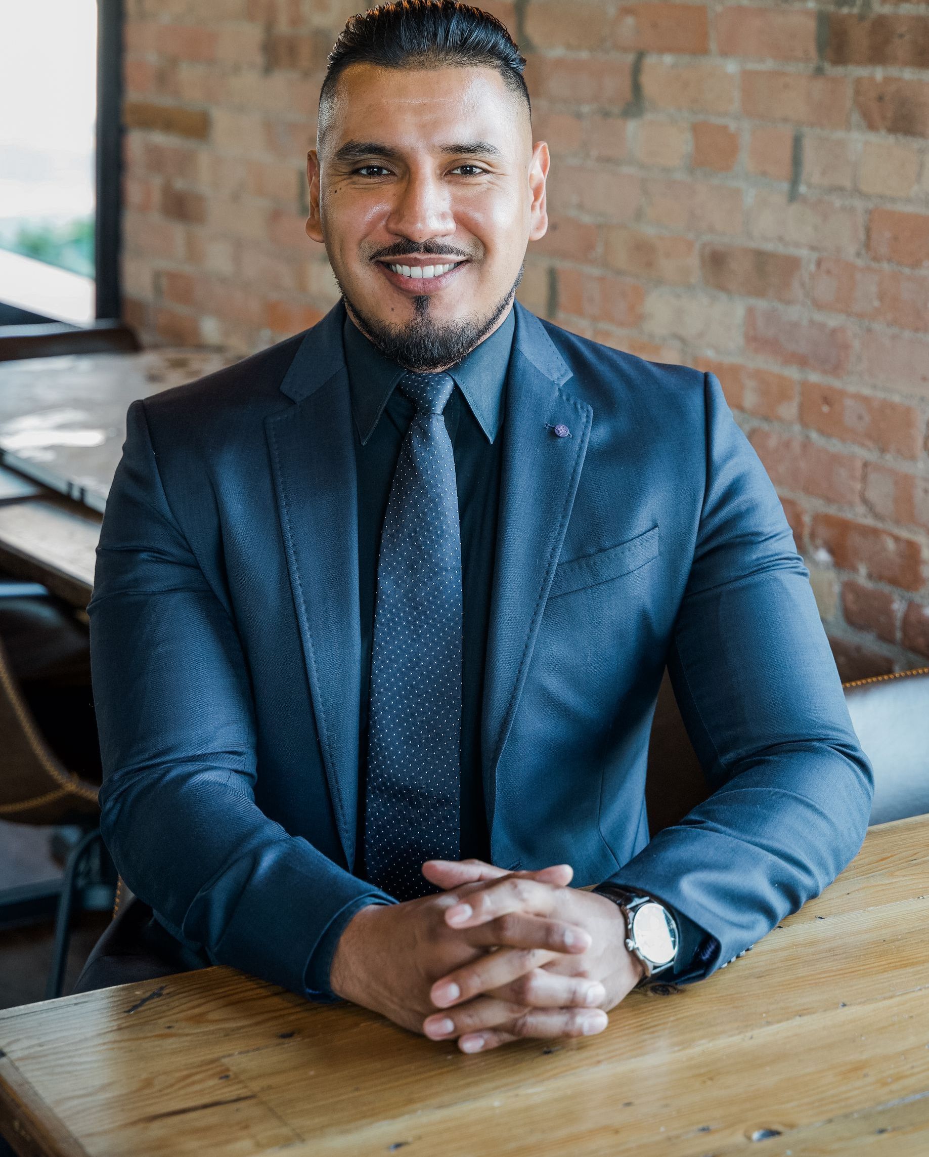 Man in blue suit seated at a wooden table in a brick-walled office, smiling with hands clasped.