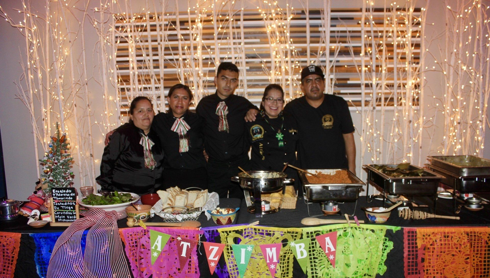 Group of five standing behind a decorated food table at a festive indoor event.