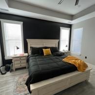 A bedroom with a black accent wall, a white bed frame, black bedding, a mustard yellow pillow, and two windows.