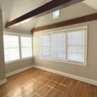 An empty room with light wood floors, beige walls, white trim, and two windows with blinds beneath exposed ceiling beams.