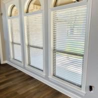 Three arched windows with white horizontal blinds installed on a light gray wall above hardwood floors.