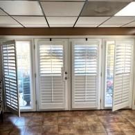 White louvered window shutters covering a double set of glass patio doors in a room with a tiled floor and drop ceiling.