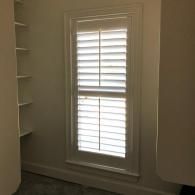 A tall, narrow window with white plantation shutters set between a light-colored wall and built-in white shelving.