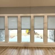 Five large windows with white roller shades look out onto a construction site with a yellow machine.