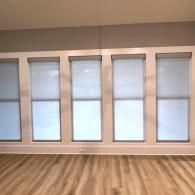 Five identical white-framed windows with translucent roller shades installed above light wood-look flooring.