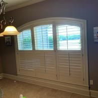 Three-panel white plantation shutters covering an arched window in a room with taupe walls.