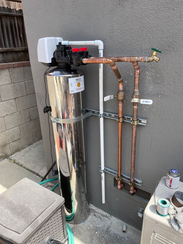 A stainless steel water filter is attached to a wall next to copper pipes.