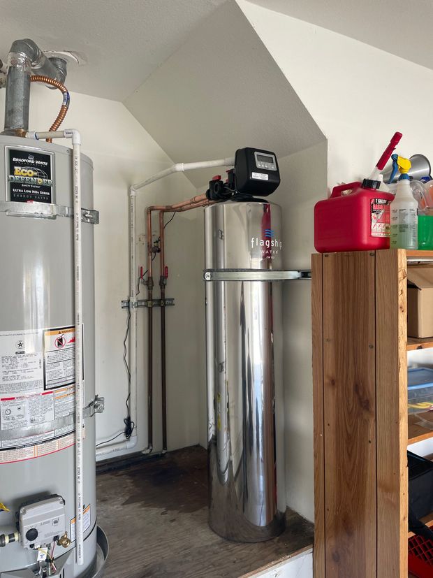 A stainless steel water heater is in a room next to a wooden shelf.