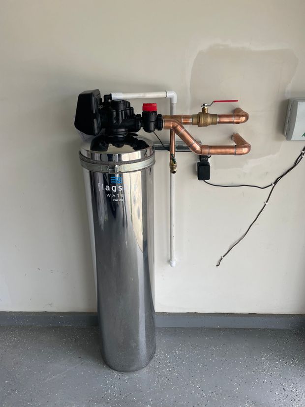 A stainless steel water filter is sitting in a garage next to copper pipes.