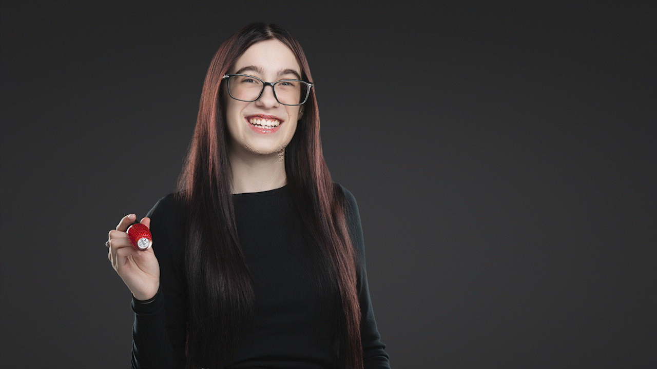 Smiling woman with long dark hair and glasses holding a small red object against a dark background