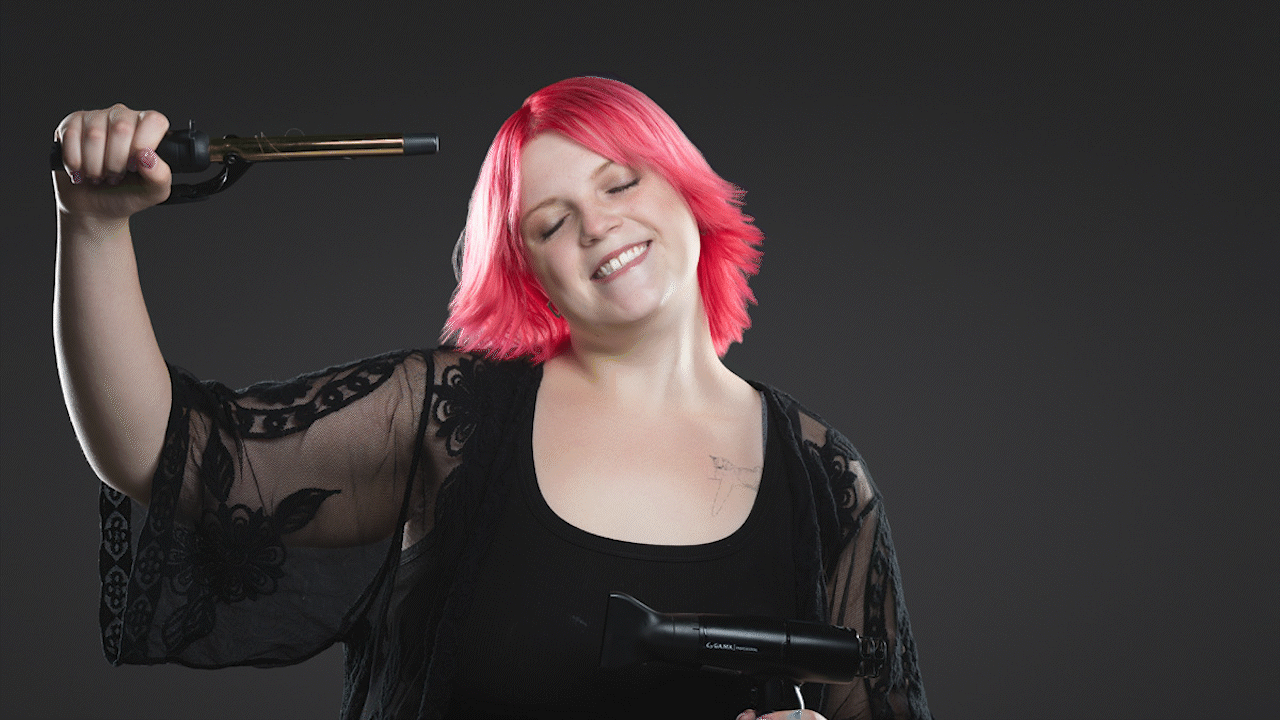 Smiling person with bright pink hair holding a black handheld prop against a dark background