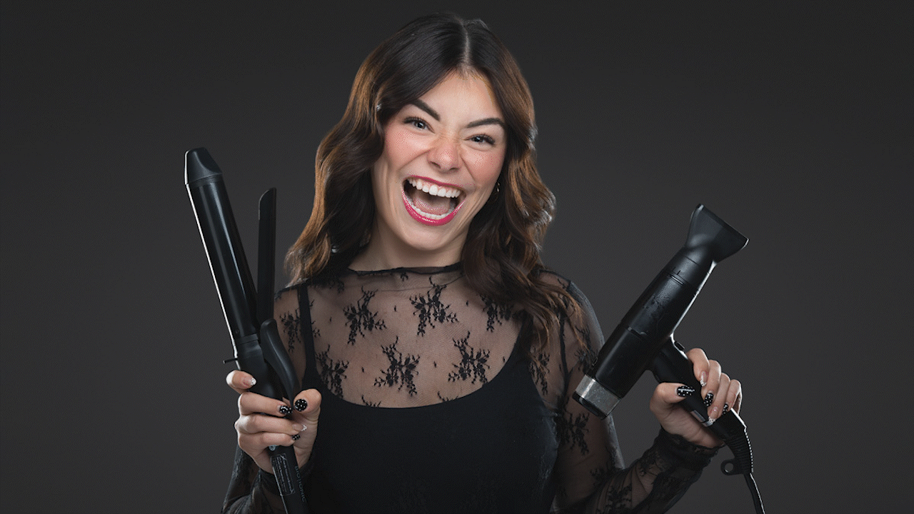 Smiling woman holding a curling iron in one hand and a hair straightener in the other against a dark background