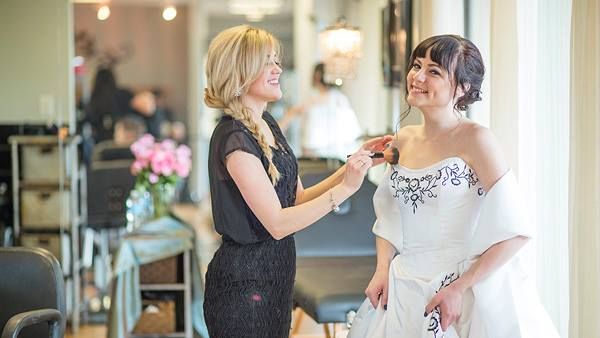 Woman adjusts necklace on smiling bride in white dress, indoors at a wedding venue.