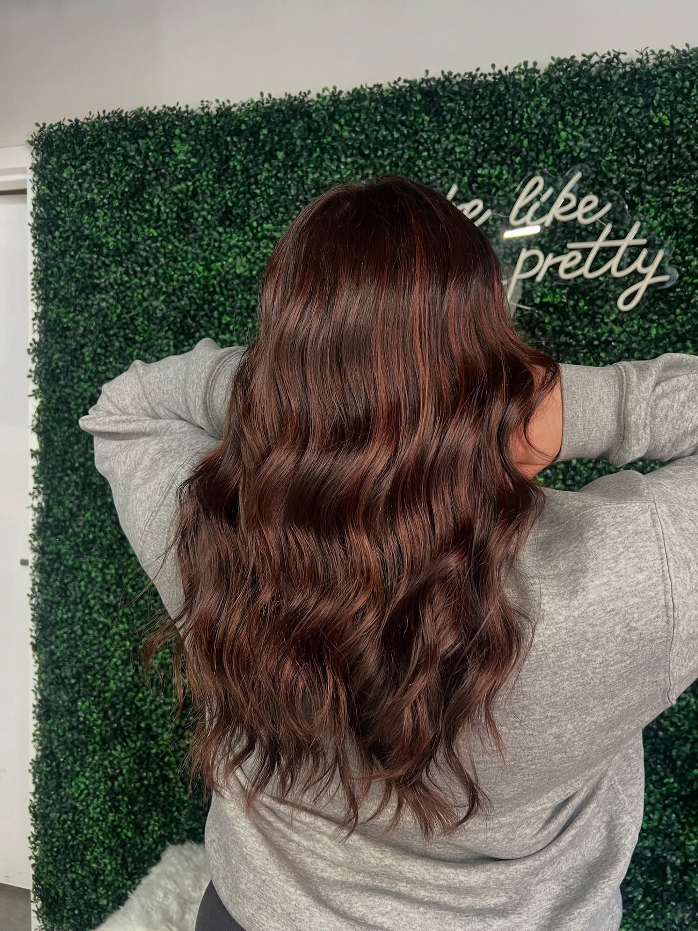 Person with wavy brown hair facing a green hedge wall with white script reading “be pretty”