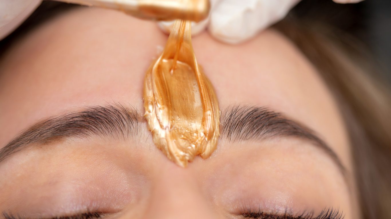 Close-up of a hand applying golden wax to a person’s eyebrow area