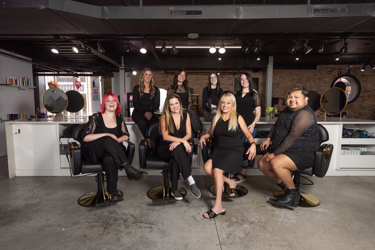Five people posing in black outfits in a tattoo studio, with mirrors and salon chairs in the background.