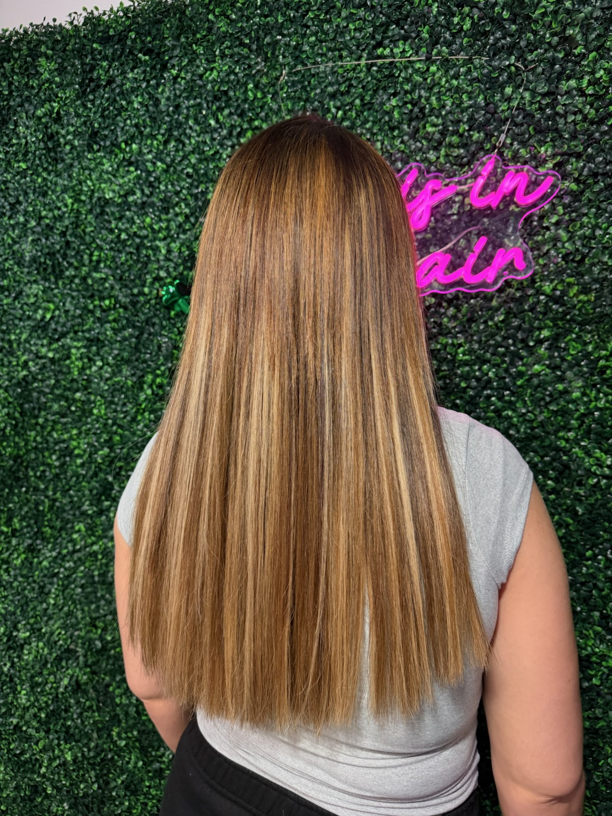 Person with long straight light brown hair stands before a green hedge wall with pink neon sign text.