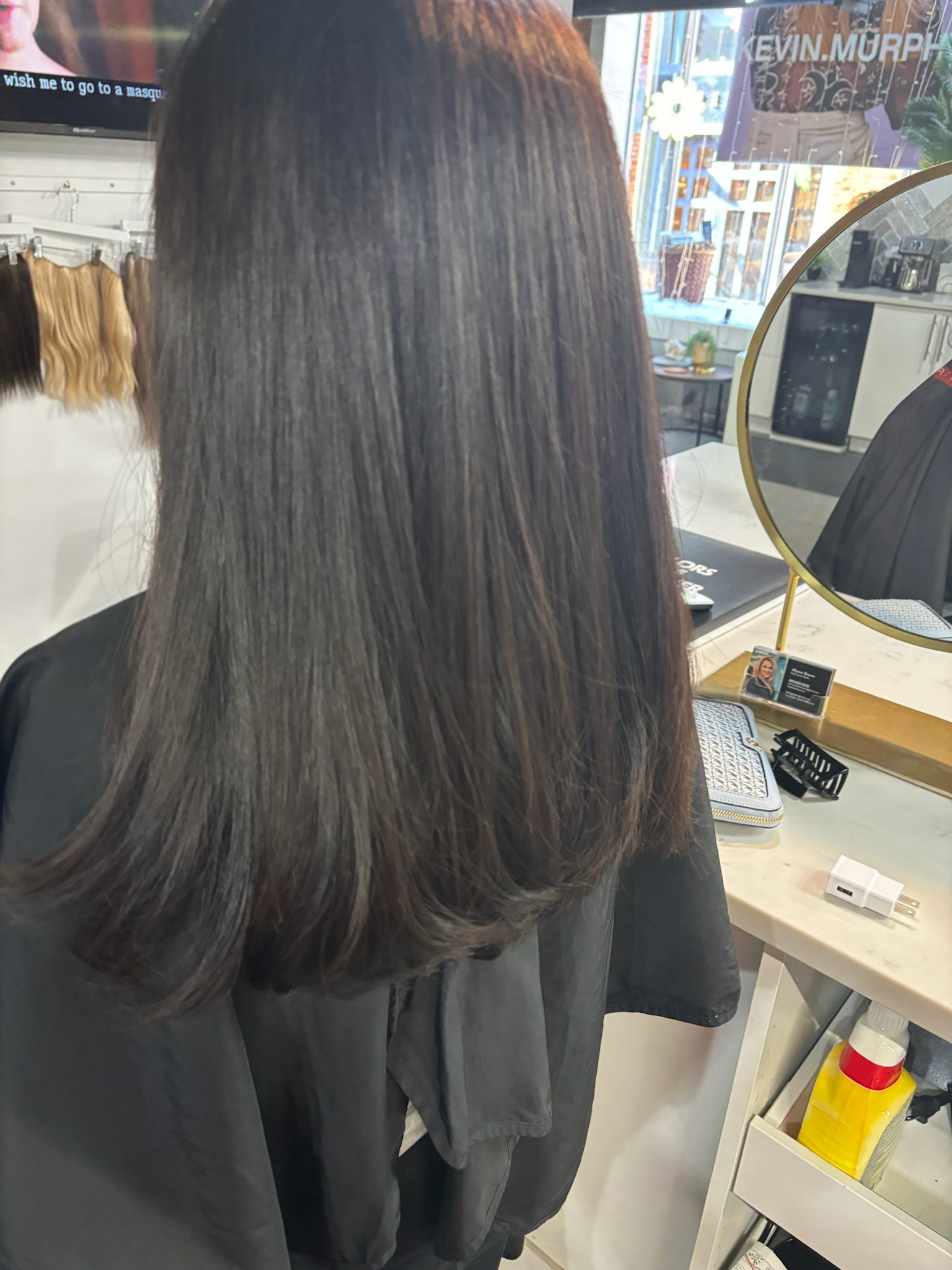 Long, straight dark hair viewed from behind in a salon mirror, with a black cape and styling tools nearby