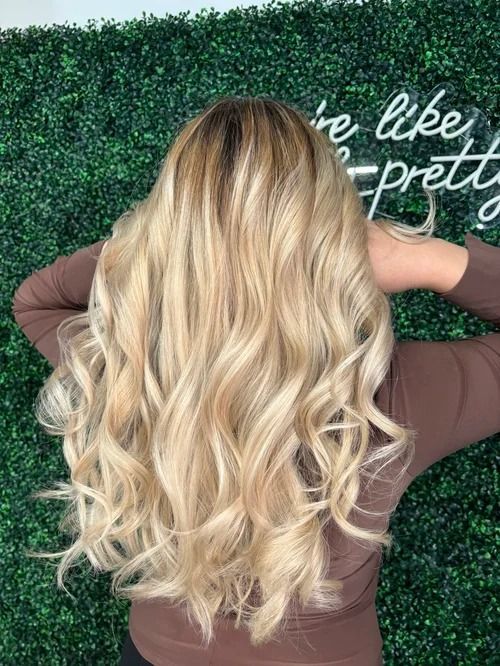 Long blonde wavy hair shown from behind against a green hedge backdrop