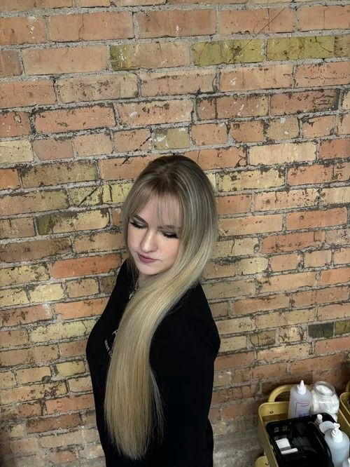 Woman with long blonde hair standing before a brick wall, wearing a black top.
