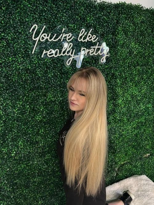 Blonde woman posing before a green hedge wall with neon sign reading “You’re like really pretty.”