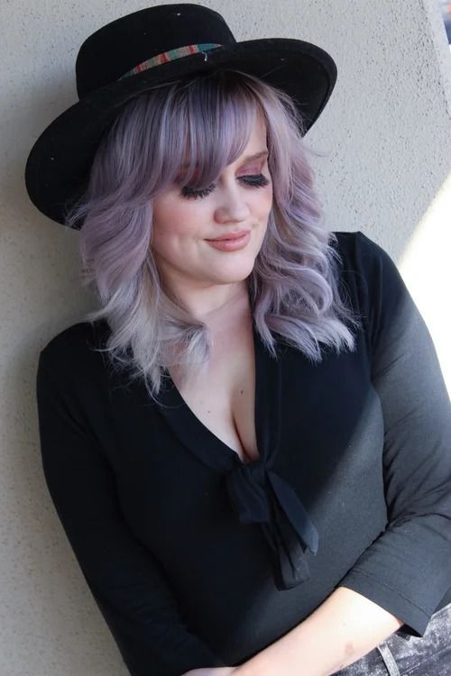 Woman in a black hat and top with silver hair, looking down against a light wall.