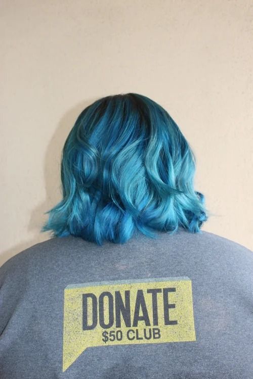 Person with blue hair wearing a gray shirt with a yellow “DONATE $50 CLUB” graphic on the back