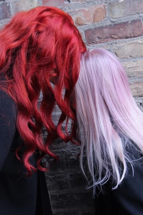 Two people with long red and pale pink hair stand with their backs to a brick wall.