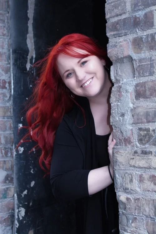 Red-haired person smiling from behind a brick wall in a dark doorway