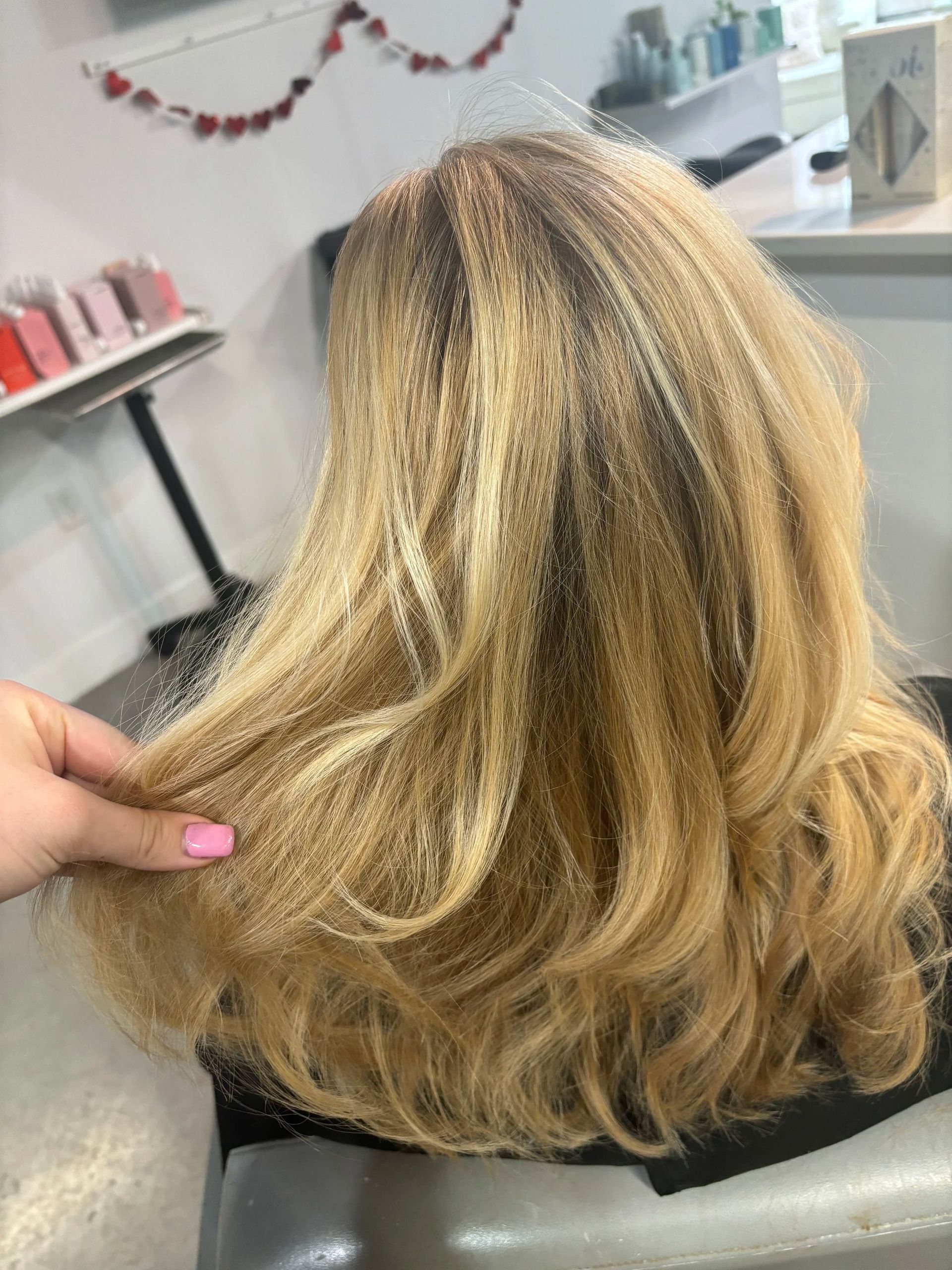 Blonde wavy hair viewed from behind in a salon, with a hand lifting the hair to show texture.