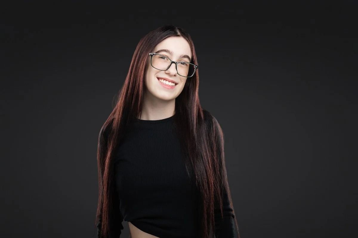 Smiling person with long dark hair and glasses, wearing a black top against a dark background