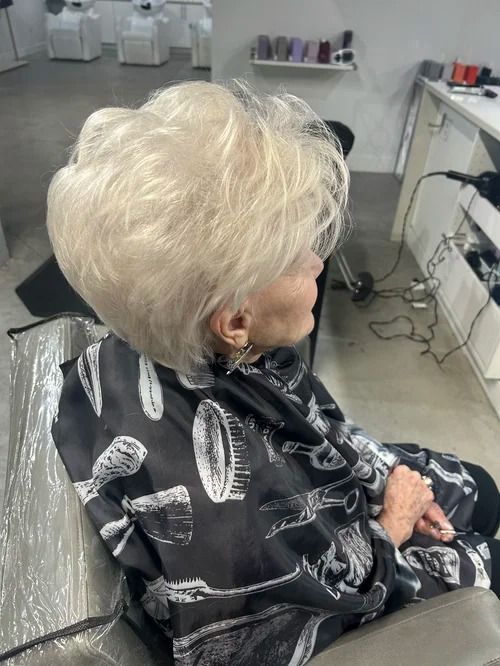 Person with short white hair seated in a salon chair, wearing a black cape with white scissors and combs.