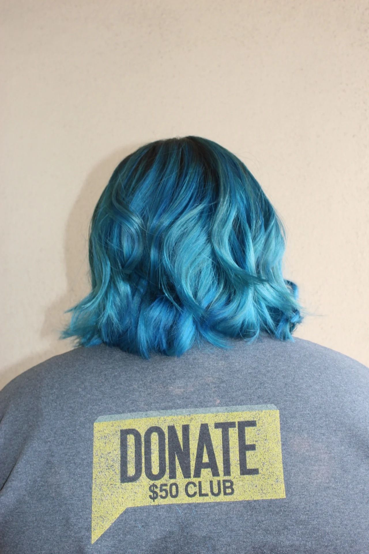 Person with bright blue hair wearing a gray shirt with a yellow “DONATE $50 CLUB” graphic on the back