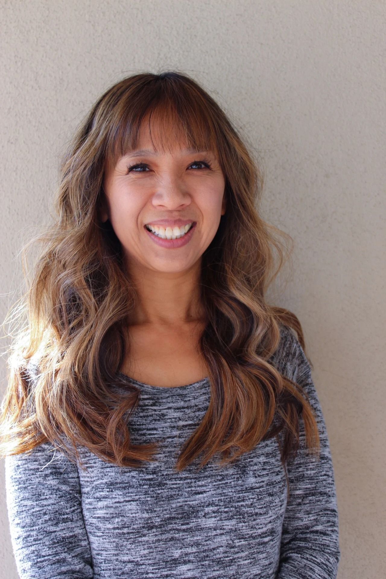 Smiling woman with long wavy hair wearing a gray sweater against a plain wall