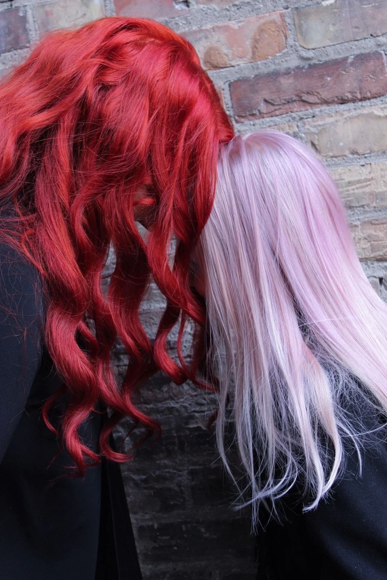 Two people with long red and lavender hair seen from behind against a brick wall.