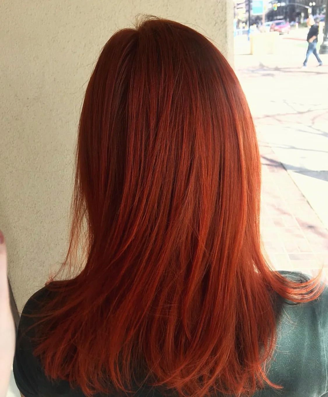 Back view of a person with straight, shoulder-length red hair standing indoors by a bright window.