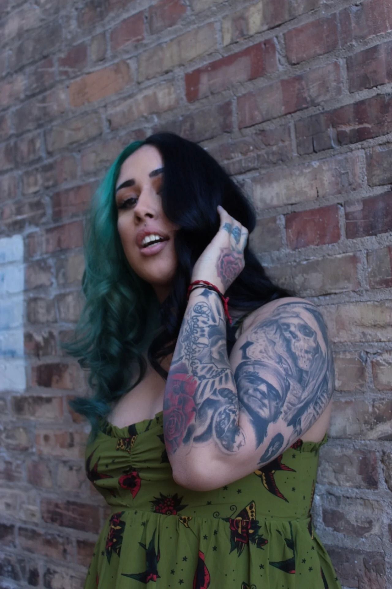 Person with teal hair and tattoos leaning against a brick wall, smiling in a green top.