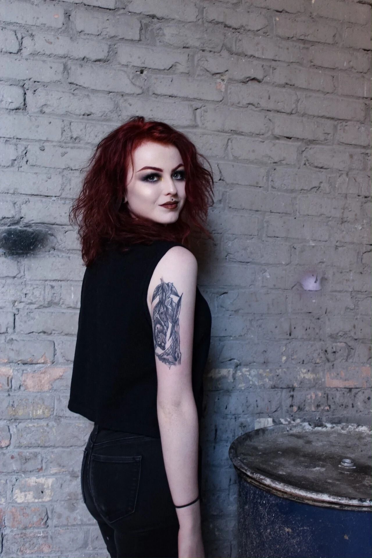 Woman with curly red hair standing against a gray brick wall, wearing black, with a tattoo on her upper arm