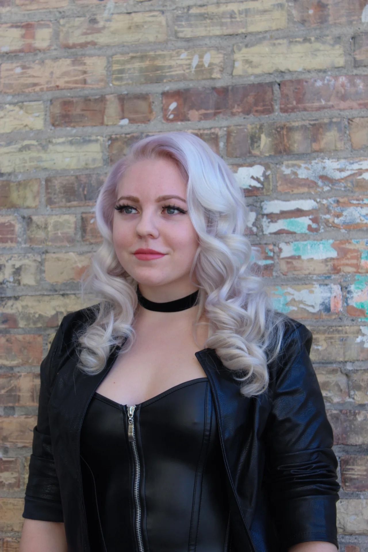 Person with silver curls in a black leather jacket and choker, standing against a brick wall.